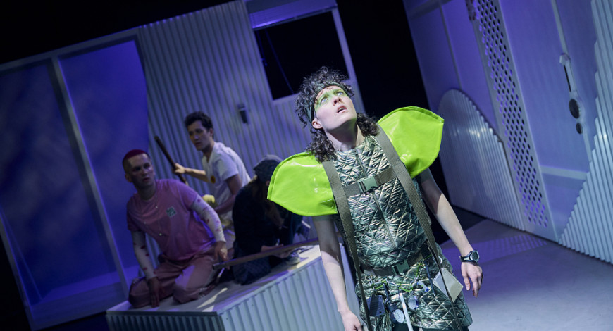 Szenenfoto aus der Inszenierung Funken. Toni Pitschmann als Twinkle schaut nach oben. Im Hintergund sitzen die anderen Darsteller*innen auf einem weißen Kasten im Bühnenbild.